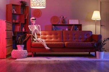 Human skeleton model with sunglasses and cup sitting on couch in living room at night