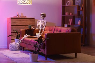 Human skeleton model with sunglasses and cup sitting on couch in living room at night