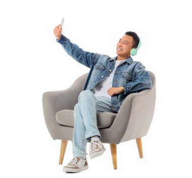 Young Asian man with headphones taking selfie in grey armchair on white background