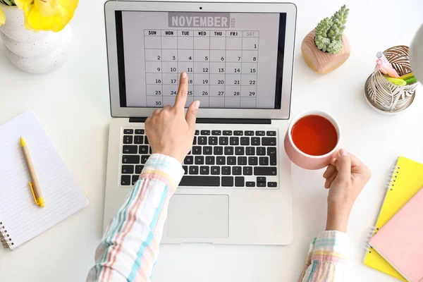 Masada takvim ve çay fincanı olan dizüstü bilgisayarı kullanan kadın eller.