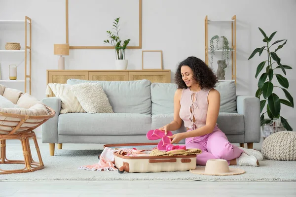 Young African-American woman packing flip-flops into suitcase for trip at home