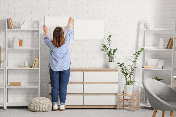 Genç bir kadın evdeki beyaz tuğla duvara resim asıyor.