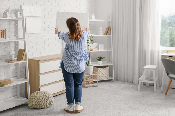 Genç bir kadın evdeki beyaz tuğla duvara resim asıyor.