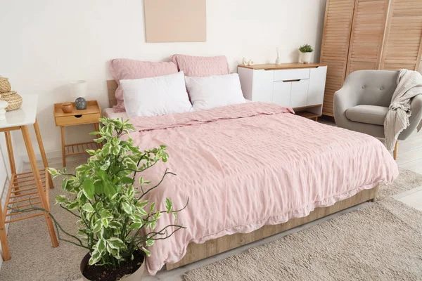 Bed with stylish pink bedding, folding screen, commode, armchair, table and houseplants in bedroom