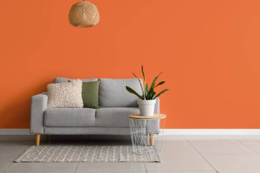 Oturma odasının iç tarafındaki turuncu duvarın yanındaki masada rahat bir kanepe ve çiçek var.