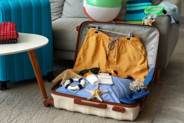 Erkek mayosu, pasaport ve plaj aksesuarlarıyla dolu bavulu kanepenin yanında aç.