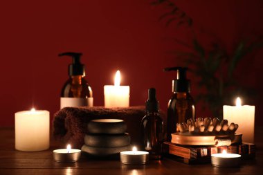 Masaj fırçası, spa taşları, kozmetik ürünleri şişeleri ve kırmızı duvarın yanındaki masada yanan mumlar.