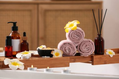 Plumeria çiçekleriyle dolu küvet tepsisi, temiz havlular, kozmetik ürünler şişeleri ve banyoda yanan mumlar, yakın çekim.