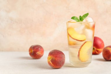 Şeftali dilimli bir bardak içecek, buz küpleri ve bej duvarlı beyaz masa üzerinde nane yaprakları.