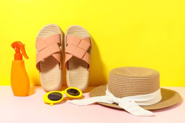 Sarı arka planda şık plaj ayakkabıları, şapka ve bir şişe güneş kremi, yakın plan.
