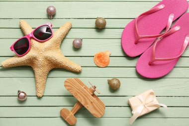 Güneş gözlüklü denizyıldızı, parmak arası terlik, tahta uçak ve yeşil ahşap arka planda Noel topları. Üst görünüm