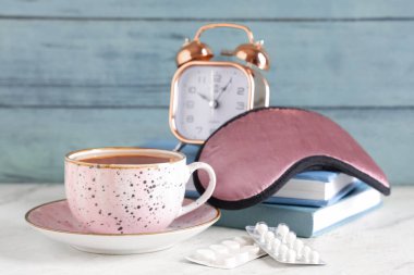 Bir fincan çay, uyku maskesi, kitaplar, haplar ve çalar saat beyaz masada.