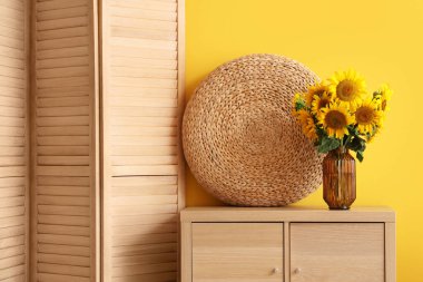 Sarı duvarın yanındaki klozette, katlı ekranlı şık bir oda, sedir ve vazo dolusu ayçiçeği buketi, yakın plan.