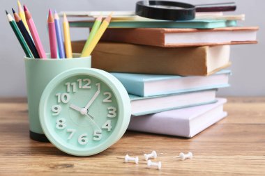 Kitap yığını, renkli kalemler, alarm saati ve ahşap masada gri duvara karşı büyüteç.