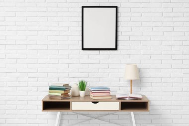 Beyaz tuğla duvarın yanındaki masada yığınla kitap, lamba ve çiçek var.