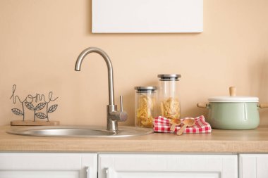 Mutfaktaki bej duvarın yanındaki tezgahın üzerinde makarna kavanozları ve pişirme kabıyla lavabo.