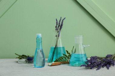 Yeşil duvarın yanındaki gri masada doğal kozmetik ürünlerini hazırlamak için malzemeler ve numunelerle dolu laboratuvar vitrinleri.