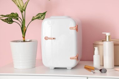 Küçük kozmetik buzdolabı, ev bitkisi ve çekmecelerin üzerinde pembe arka planda duran kozmetik çanta. Yakın çekim.