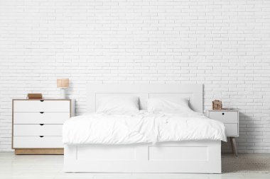 Büyük yataklı ve beyaz tuğla duvarın yanında çekmeceleri olan açık renkli bir yatak odası.