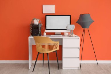 Ofiste turuncu duvarın yanında boş bilgisayar ve lamba bulunan mucit işyeri.