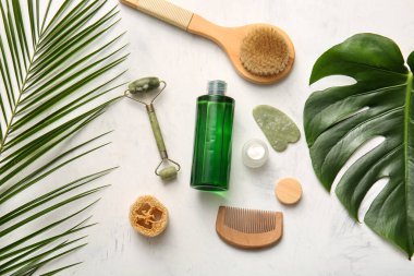 Tropikal yapraklı spa kompozisyonu, yüz masajı araçları, kavanoz ve beyaz arka planda bir şişe kozmetik ürünü, yakın plan