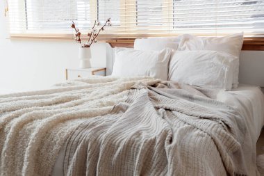 Yatak odasındaki pencerenin yanındaki komodinin üzerinde pamuk çiçekleri olan rahat bir yatak ve vazo.