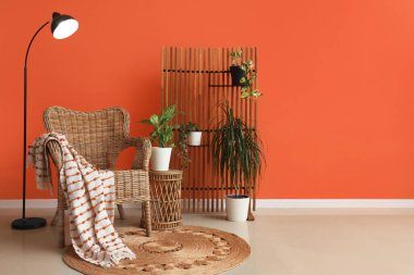 Hasır koltuğu, çiçekleri ve turuncu duvarın yanında standart lambası olan şık bir oturma odası.