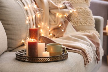 Farklı yanan mumlarla dolu bir tepsi ve oturma odasındaki koltukta bir fincan çay, yakın çekim.