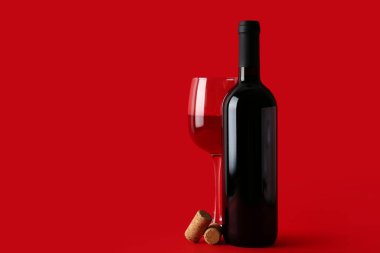 Kırmızı arka planda şişe, bir bardak şarap ve mantar.