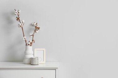 Oturma odasındaki beyaz duvarın yanındaki çekmecelerde pamuk çiçekli fotoğraf çerçevesi, fincan ve vazo.