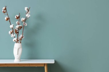 Oturma odasındaki yeşil duvarın yanındaki masada pamuk çiçekli vazo, yakın plan.