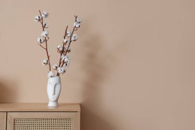 Oturma odasındaki bej duvarın yanındaki çekmecelerin üzerinde pamuk çiçekler olan vazo, yakın plan.