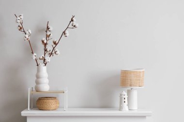 İçinde pamuk çiçekler, lamba ve biblo olan vazo, oturma odasındaki beyaz duvarın yanındaki çekmecelerin yanında, yakın plan.