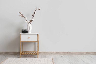 Oturma odasındaki beyaz duvarın yanındaki masada pamuk çiçeklerle fotoğraf makinesi ve vazo.