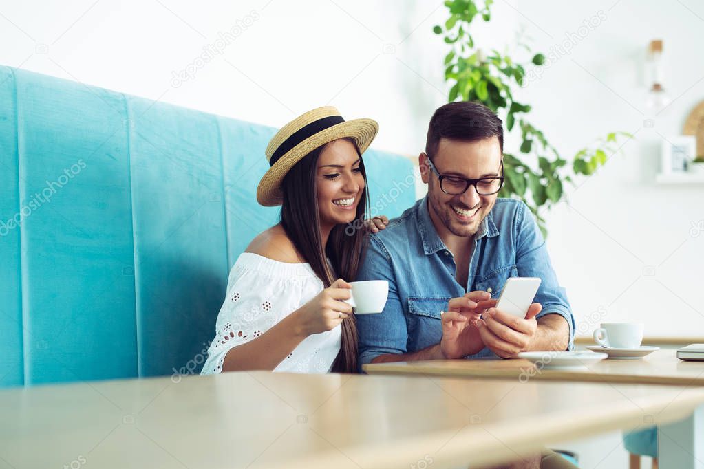 Happy young couple sitting at the cafe table drinking coffee and looking at mobile phone