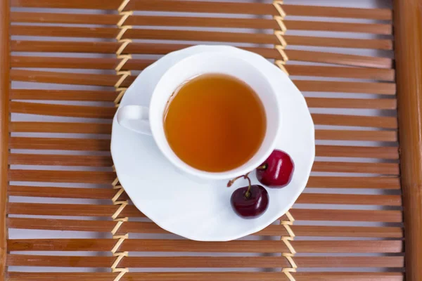 A cup of tea and a sweet cherry on a saucer.