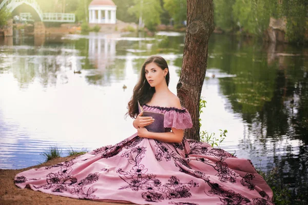 A beautiful young woman in a 19th-century dress sits with a book by the pond. Romantic girl in the park.