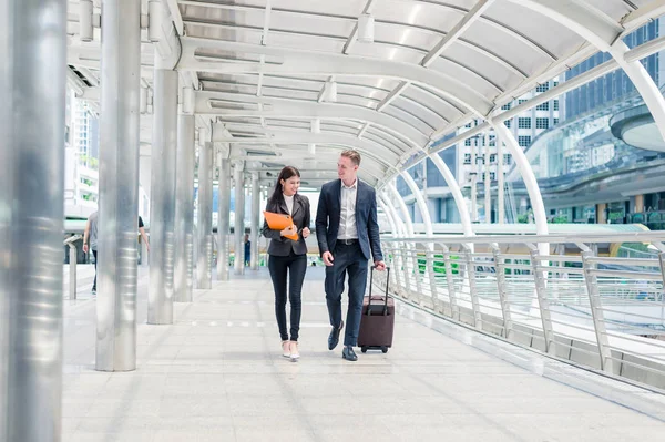 Hombre de negocios con equipaje y mujer de negocios con archivo de documentos a mano caminan juntos en la ciudad, concepto de viaje de negocios — Foto de Stock
