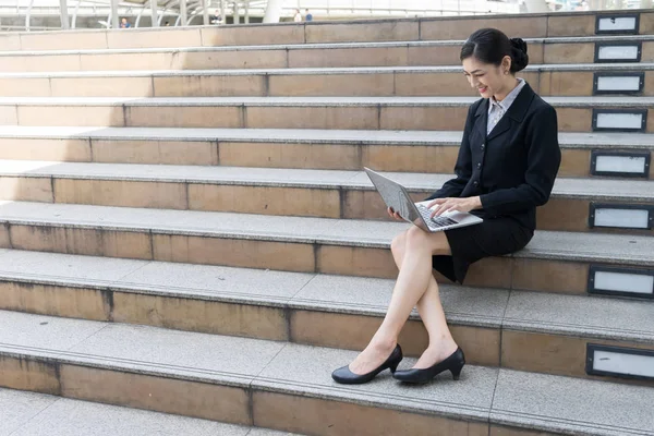 Asiatisk forretningskvinne smiler og sitter i trappa og skriver ord inn i en bærbar maskin. – stockfoto