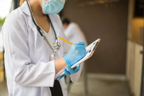 Imagem de médico mulher em casaco branco com estetoscópio e luva azul escrever relatório por lápis em um caderno sobre fundo borrão do hospital . Imagens De Bancos De Imagens Sem Royalties