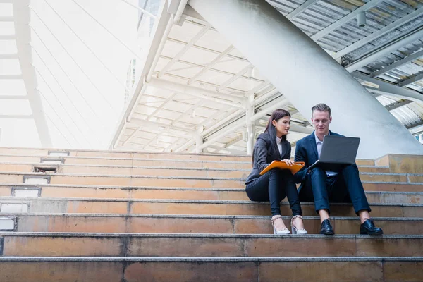 La mujer de negocios asiática y el hombre de negocios caucásico se sientan en la escalera y sostienen un papel del informe financiero con el ordenador portátil. concepto de informe de venta y plan de marketing con espacio de copia para texto . — Foto de Stock