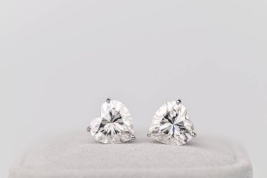 Kalp şeklinde iki elmas küpe. Küpeler beyaz ve beyaz bir yüzeyde oturuyorlar.