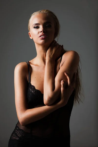 Portret van blonde jonge vrouw met mooie make-up en kapsel in een zwart kanten bodysuit staat op grijze achtergrond. Fashion model poseert in elegante kleding in de studio — Stockfoto