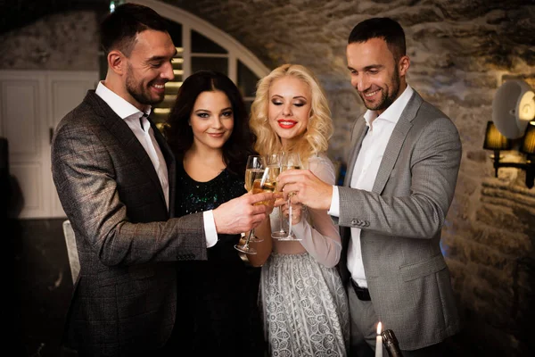 Fête, vacances avec des amis concept. Quatre personnes avec des verres de champagne célébrant et grillant au restaurant. Deux hommes et deux femmes dans des vêtements de soirée élégants, costumes et robes de nuit à l'intérieur — Photo