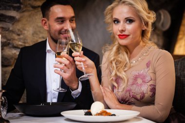 Romantizm ve flört konsepti. Şampanya bardaklı genç bir çift restoranda kutlama yapıyor ve kadeh kaldırıyor. İki kadın ve erkek şık bir gece elbisesi ve romantik bir akşam yemeği.