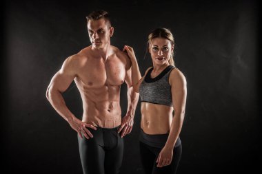 Spor, spor ve sağlıklı yaşam tarzı konsepti. İki atletik erkek ve kadın eğitimli vücutlarını siyah arka planda gösteriyorlar. İki vücut geliştirme modeli ayakta duruyor ve sıkı kasları gösteriyor..