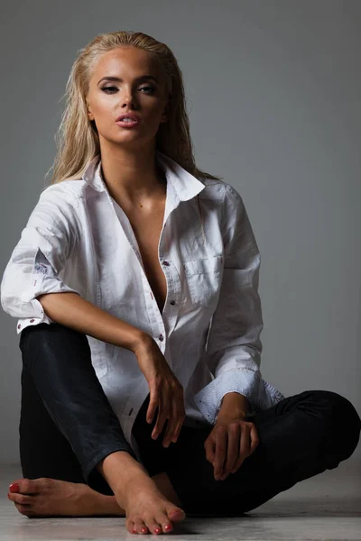 Retrato de mulher jovem loira com bela maquiagem e penteado em camisa branca senta-se no fundo cinza. Modelo de moda posa em roupas elegantes em estúdio — Fotografia de Stock