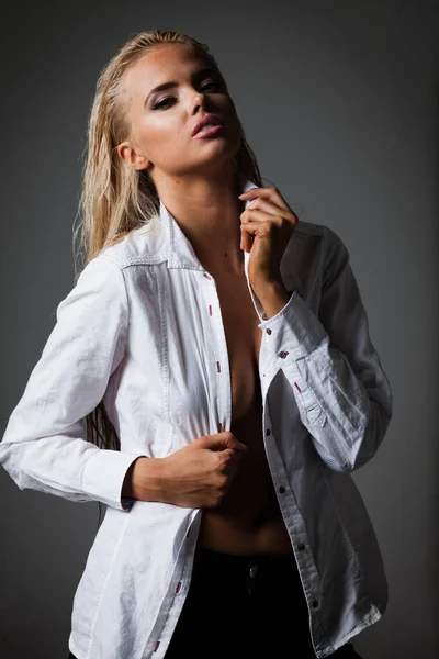 Portrait de jeune femme blonde avec un beau maquillage et coiffure en chemise blanche se dresse sur fond gris. Modèle de mode pose dans des vêtements élégants en studio — Photo