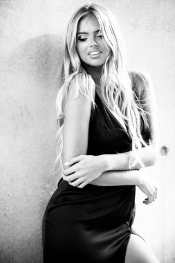 Gri duvarın yanında duran siyah elbiseli, güzel makyajlı, sarışın genç bir kadın. Şehir ortamında şık kıyafetlerle poz veren bir manken. Siyah ve beyaz, tek renkli.
