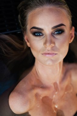 Rahatlama ve spa konsepti. Siyah mayo giymiş seksi, modaya uygun bir kadının yakın plan portresi. Islak saçlı ve makyajlı bir kadın, güneş ışığında yüzme havuzunda poz verir.